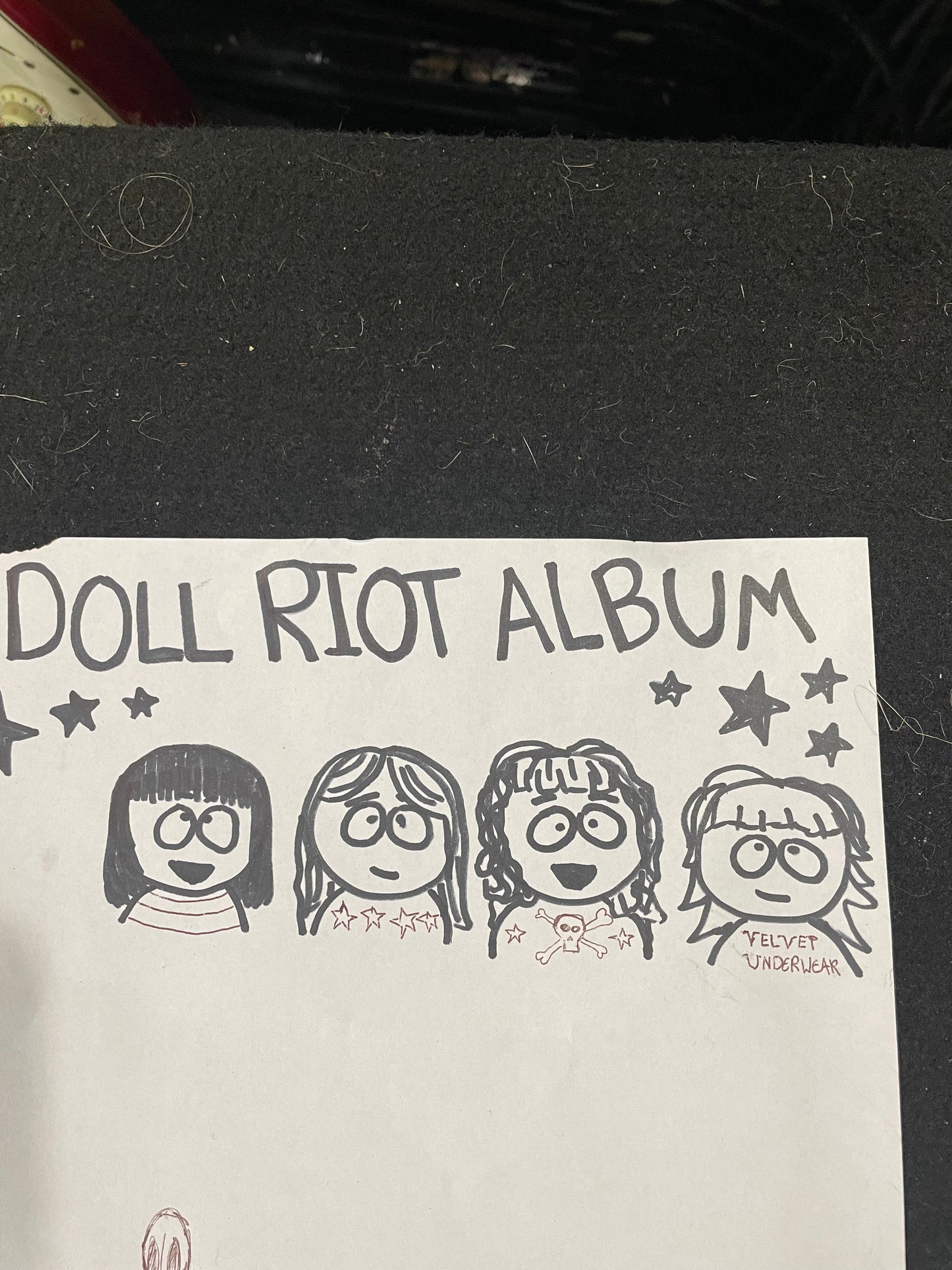 A handmade paper album cover featuring the text 'DOLL RIOT ALBUM' and four hand-drawn cartoon girl faces in black ink, placed on a dark textured surface.