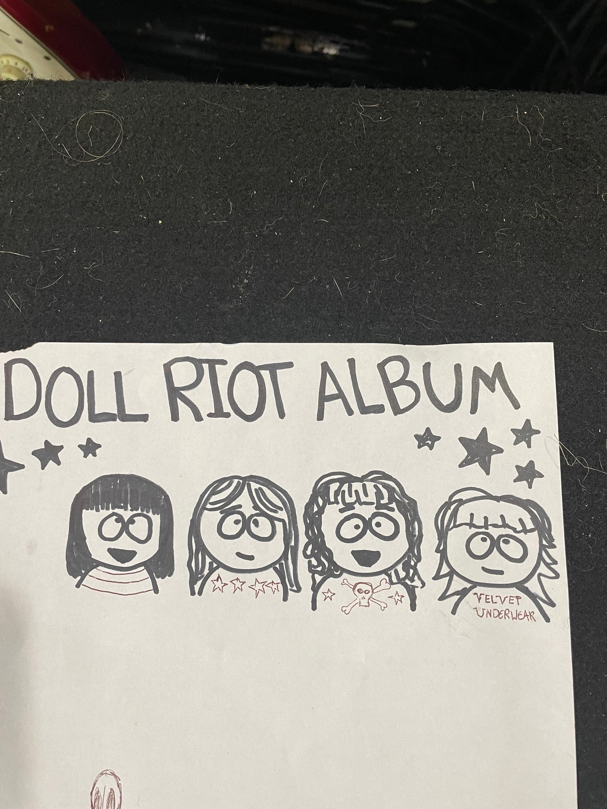 A handmade paper album cover featuring the text 'DOLL RIOT ALBUM' and four hand-drawn cartoon girl faces in black ink, placed on a dark textured surface.